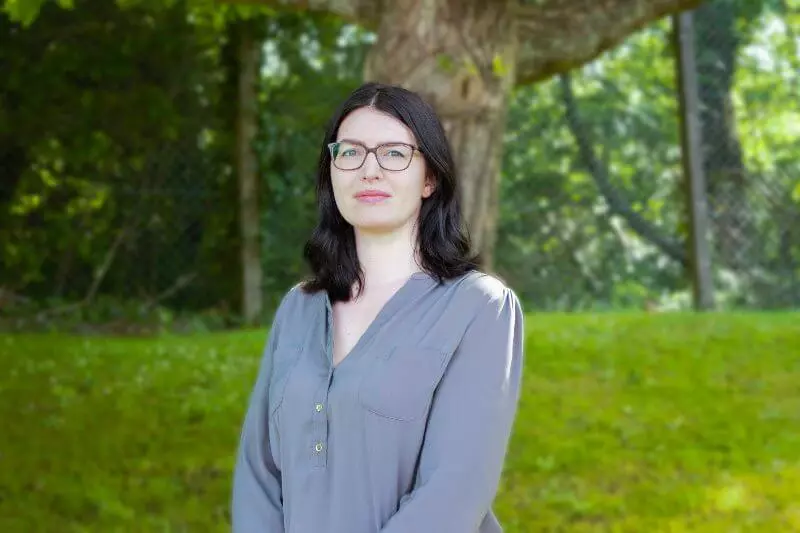 A woman in a grey top stands next to grass and a tree.