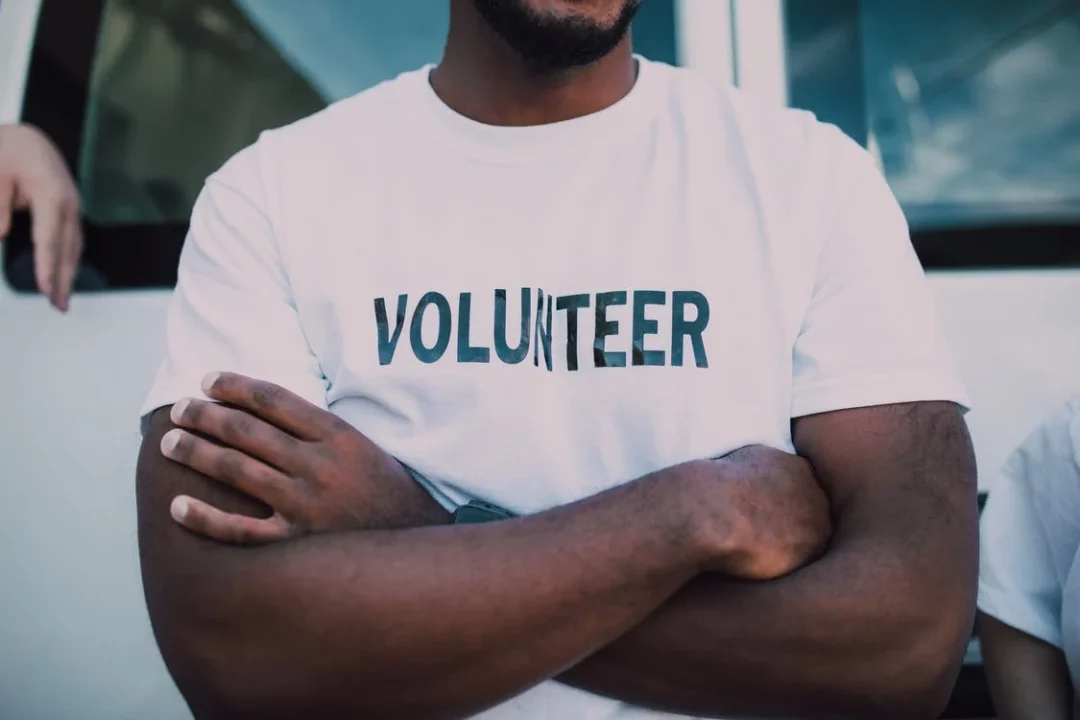 A man is standing with his arms crossed. His white t-shirt says 'volunteer.'