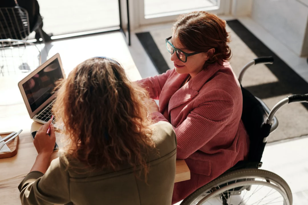 A person using a wheel chair is sat at a desk and talking to a colleague. There is a laptop in front of them.