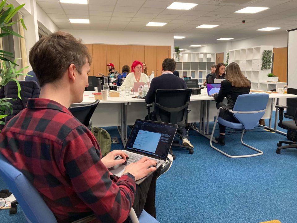 A man is sat down and working on a laptop, while others are sat at a table behind him and talking at their laptops.